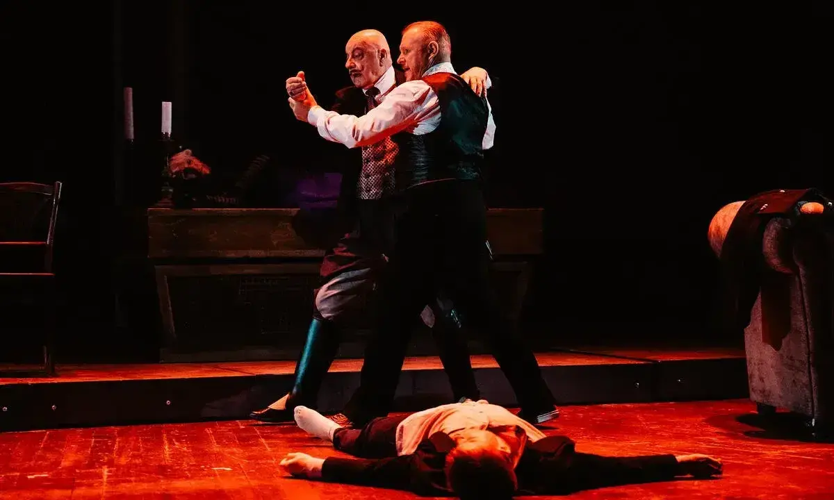 Dwóch mężczyzn tańczy tango na scenie, trzeci leży na podłodze.