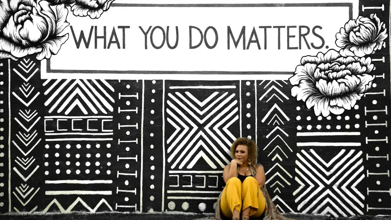 Kobieta siedzi przed muralem z napisem "WHAT YOU DO MATTERS". To inspirujący dialog motywujący.