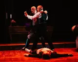 Dwóch mężczyzn tańczy tango na scenie, trzeci leży na podłodze.