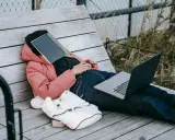 Osoba w różowej kurtce leży na ławce, z zeszytem na twarzy. Obok leży laptop i plecak. Czas na odpoczynek i dodanie nowych pomysłów.