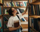 Młoda kobieta w okularach wybiera książkę z półki w bibliotece, trzymając inne w ręku.