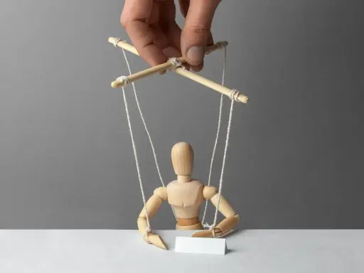 Drewniany pacynka z pustą kartką, sterowana przez rękę. Symbolizuje manipulację językową i kontrolę nad słowami.
