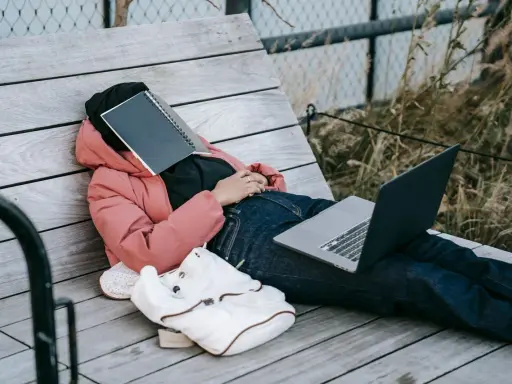Osoba w różowej kurtce leży na ławce, z zeszytem na twarzy. Obok leży laptop i plecak. Czas na odpoczynek i dodanie nowych pomysłów.