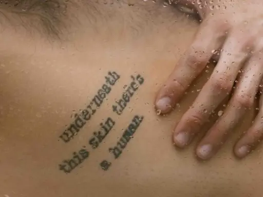 Tatuaż z cytatem "Underneath this skin there's a human" na skórze, z kroplami wody.