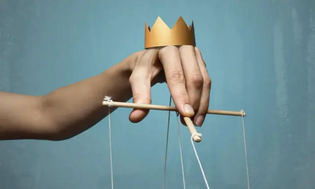Dłoń z papierową koroną na palcach, sterująca marionetką. Symbol władzy i despoty.