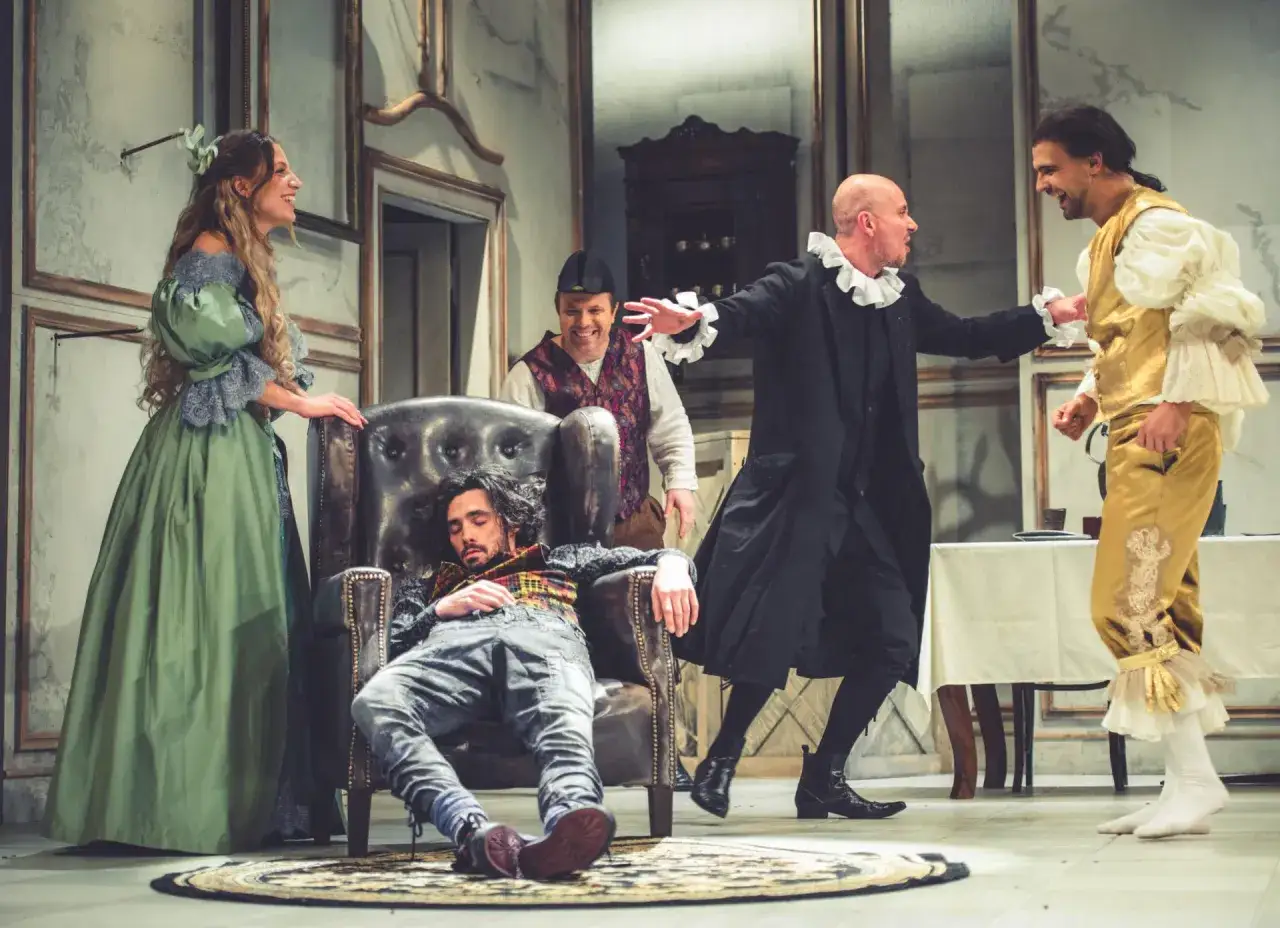 Scena teatralna: skąpiec epoka. Mężczyzna w fotelu, obok kobieta w zielonej sukni, dwaj panowie w strojach z epoki rozmawiają.