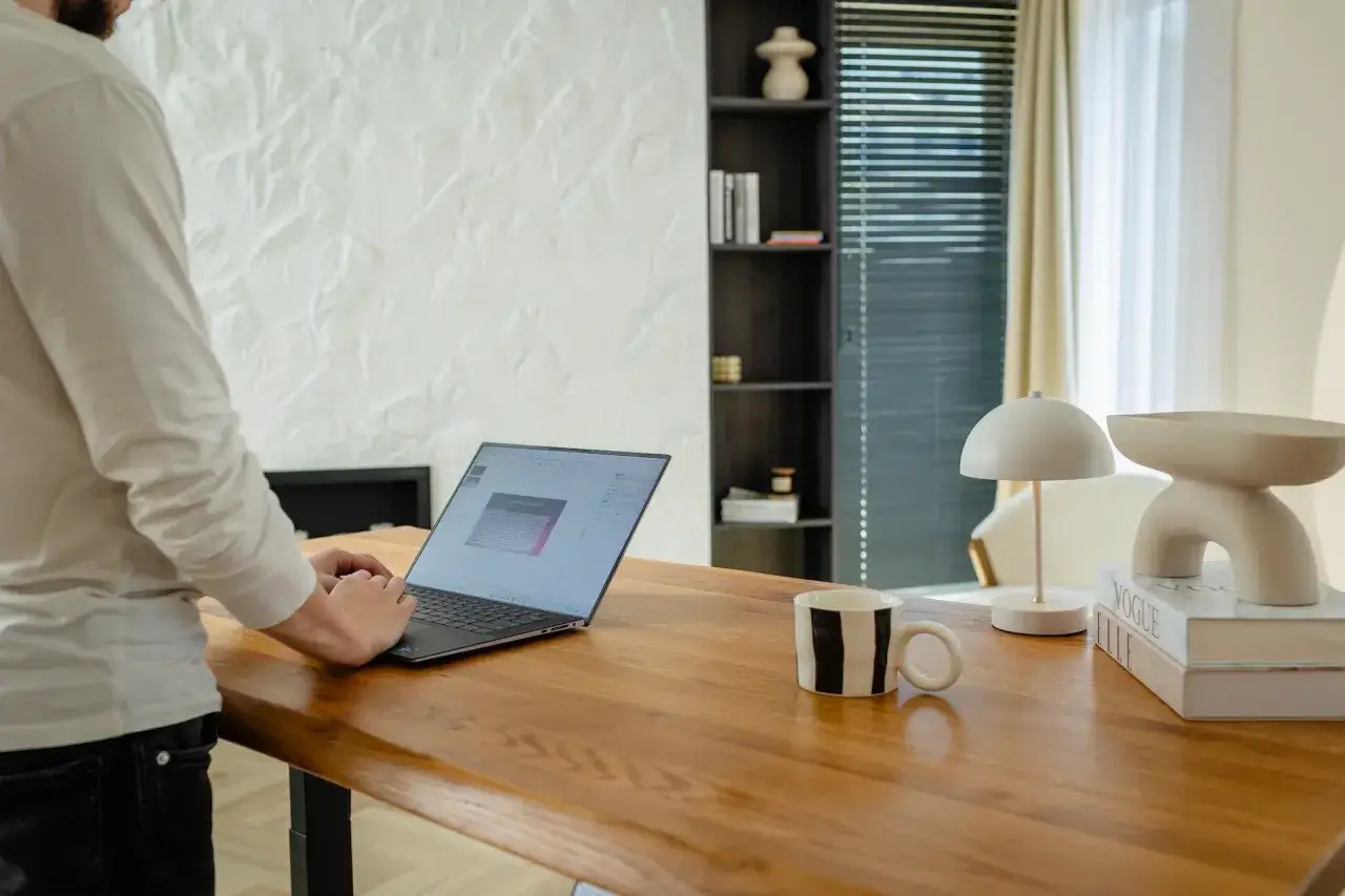 Mężczyzna pracuje przy laptopie na drewnianym biurku. Czy to jego codzienna rutyna? Obok kubek, lampka i książki.